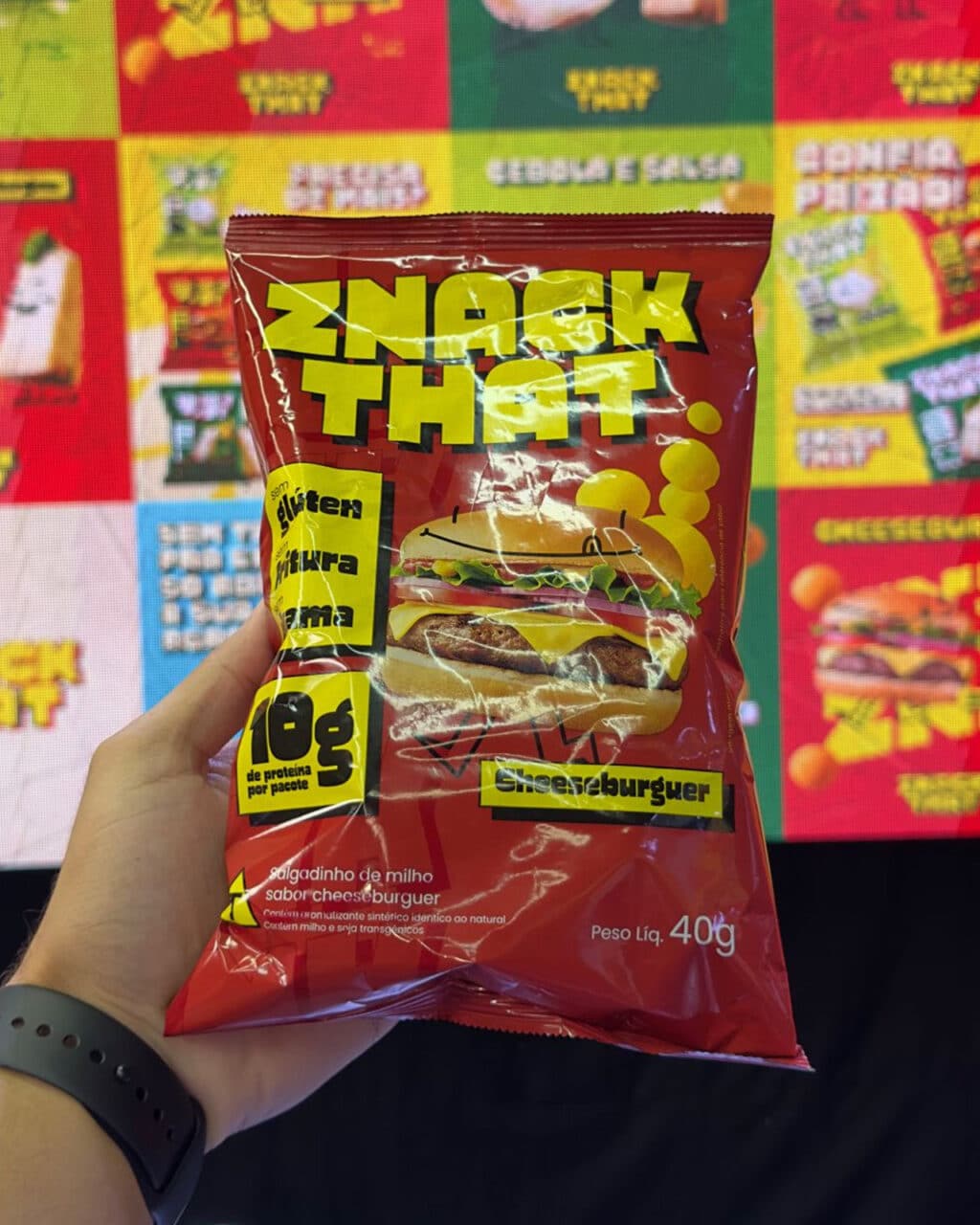 Uma mão segurando um pacote de batatas fritas com sabor de cheeseburger da marca 'Snack That' contra um fundo colorido, exibindo diversos produtos de salgadinho.