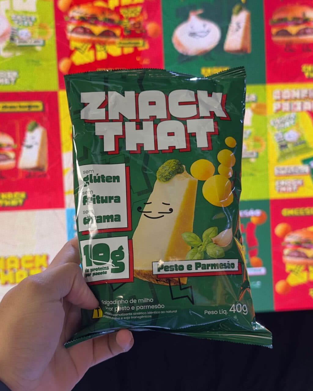 Uma mão segurando um pacote de salgadinhos verde com a etiqueta 'ZNACK THAT', nos sabores pesto e parmesão, com texto indicando que é sem glúten e contém 10g de proteína por pacote. Ao fundo, são visíveis ilustrações coloridas relacionadas a salgadinhos.