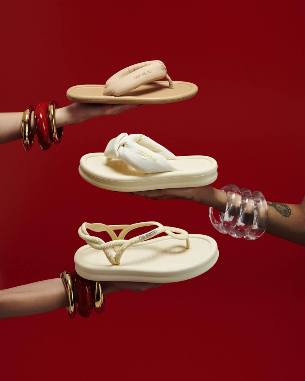 Pés com sandálias Havaianas brancas estilo “O Diabo Veste Prada” sobre fundo vermelho, em mãos com pulseiras douradas
