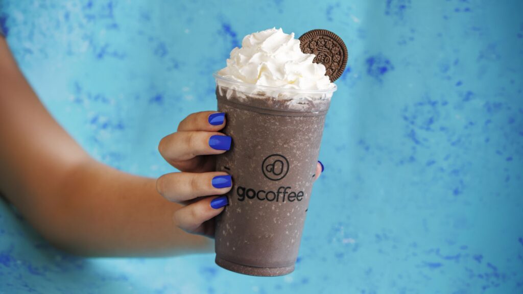 Copo de Frappe Oreo Go Coffee com chantilly em fundo azul, sendo segurado por uma pessoa
