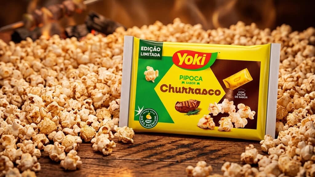 Embalagem de pipoca de micro-ondas Yoki edição limitada sabor Churrasco, cercada por pipocas na mesa.
