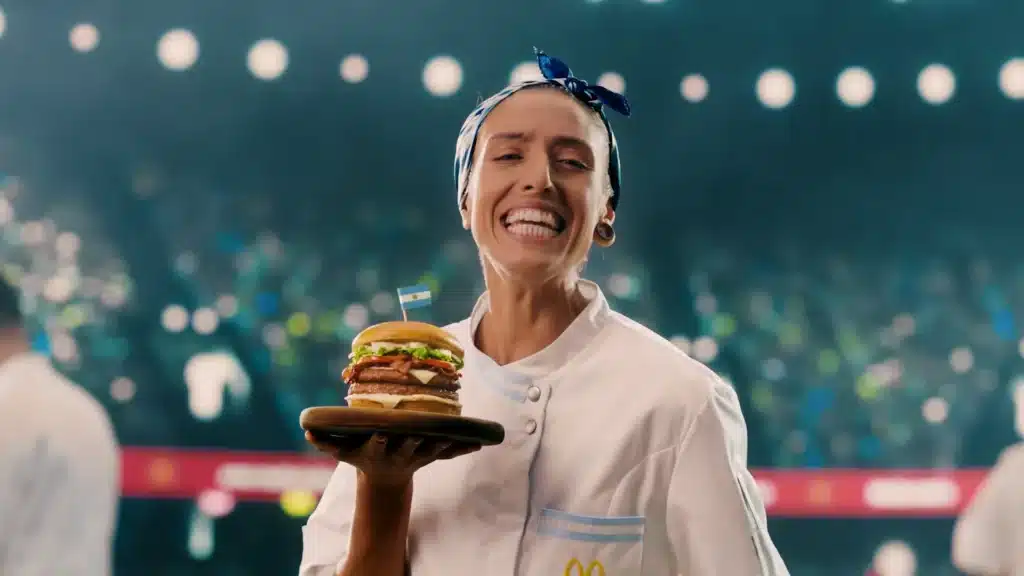 Uma cozinheira sorridente segurando um hambúrguer em um prato, vestindo uniforme branco e faixa azul na cabeça, com um fundo de estádio iluminado.