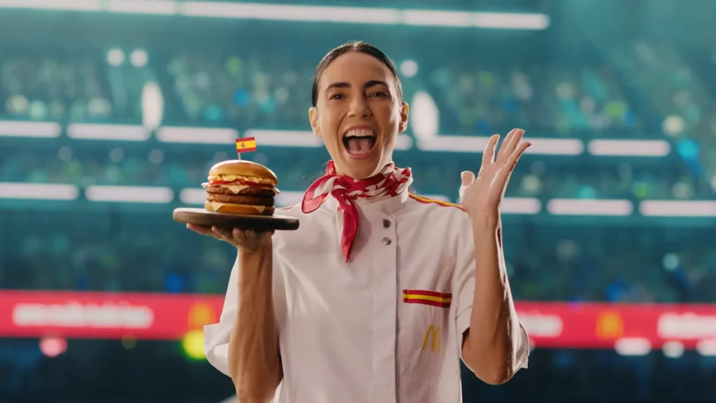 Uma mulher sorridente, vestindo um uniforme de cozinheira, segura um hambúrguer em um prato com uma bandeira da Espanha no topo, em um ambiente de estádio.