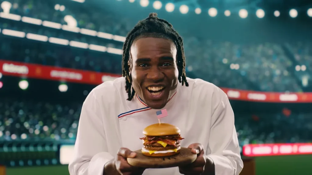 Um cozinheiro sorridente segurando um hambúrguer em um cenário de estádio, com torcedores ao fundo.