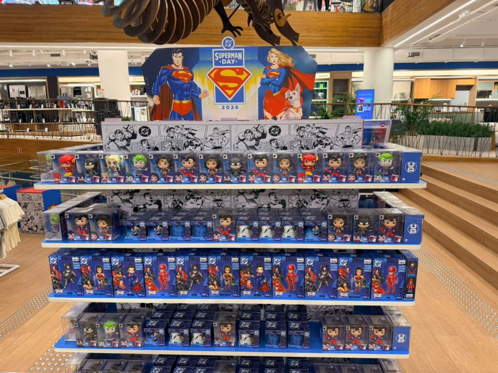Expositor com bonecos e caixas do Superman Day em loja, com cartaz do evento ao fundo