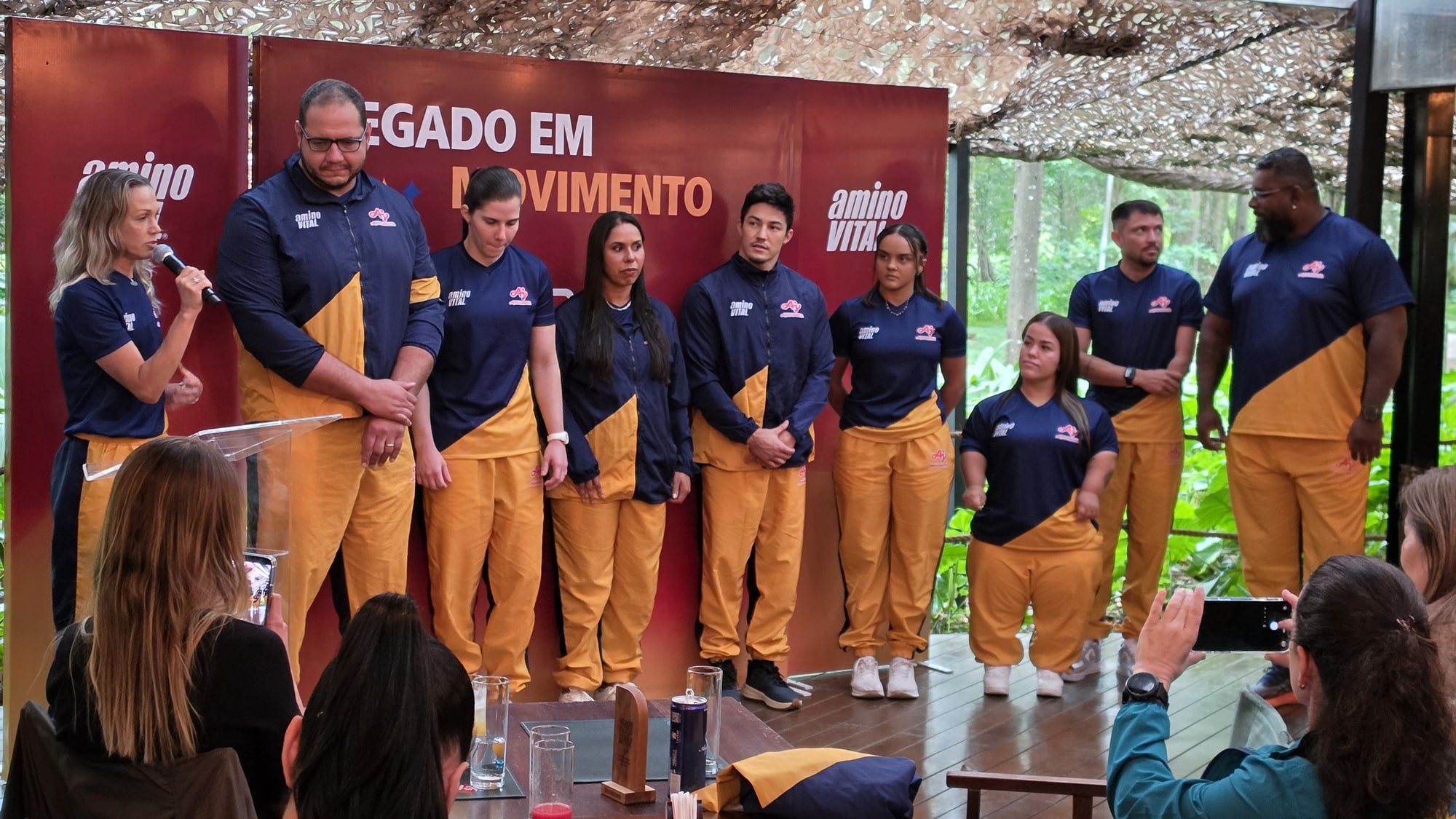 Membros da equipe com uniformes esportivos combinando em azul-marinho e amarelo, em fila durante uma apresentação ou evento, enquanto membros da plateia tiram fotos.