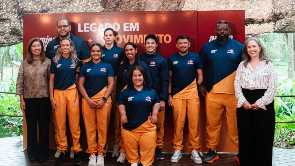 Equipe uniformizada com camisetas e calças laranja e azul em frente a um banner “Legado em Movimento”.