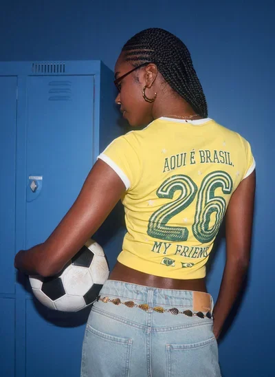 Modelo com mini-tee amarela do Guaraná Antarctica, estampa “Aqui é Brasil 26”, segurando bola perto de armário azul.