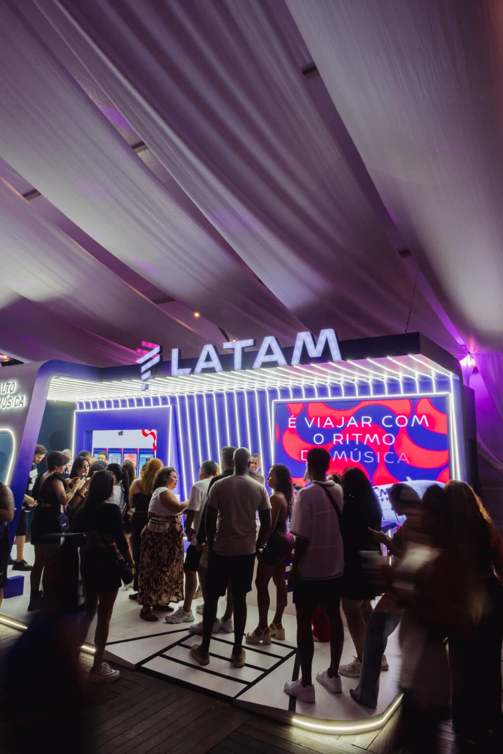 Pessoas em fila no estande LATAM TMNR, em ambiente de evento com iluminação roxa e painel de divulgação ao fundo.