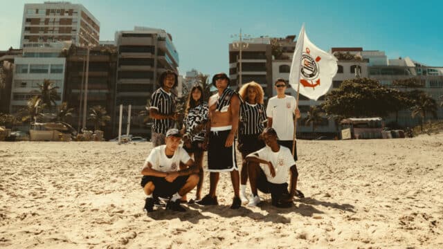 Torcida do Corinthians com bandeira em praia, em destaque roupas listradas, celebrando a invasão corinthiana de 1976