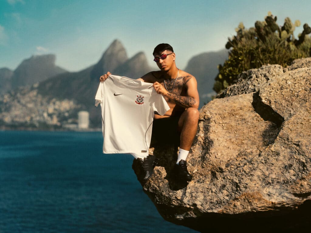 Atleta com a camisa do Corinthians branca, posando em cenário costeiro com montanhas ao fundo.