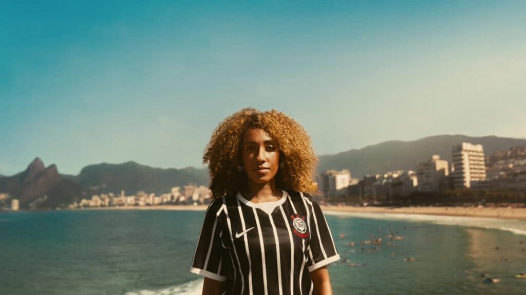 Modelo com cabelo afro veste camisa do Corinthians listrada preta e branca, em frente à praia com prédios ao fundo.