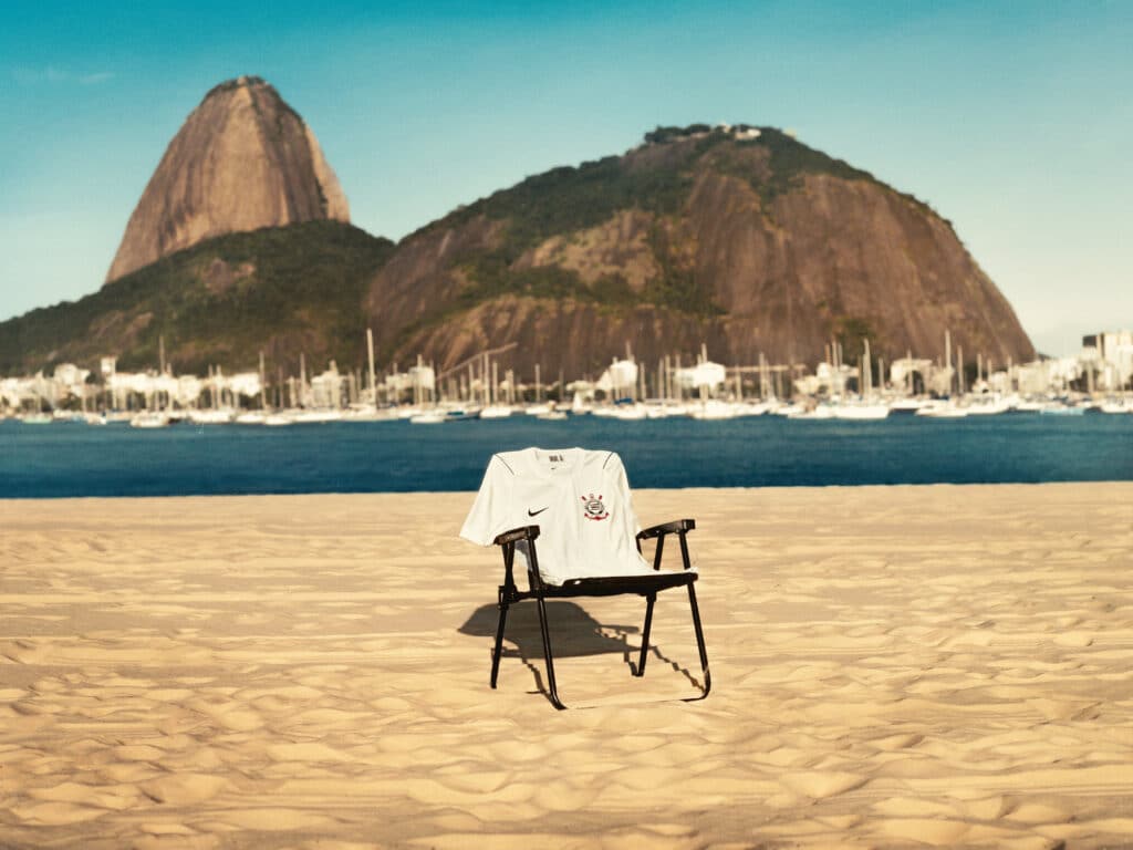 Camisa do Corinthians em fundo de praia com montanhas e cadeiras ao estilo cenário da coleção Nike Invasão 1976.