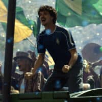 Jovem celebra no palco em evento com bandeiras do Brasil e iluminação festiva, cenário da campanha Volkswagen “Sonhos” para a Copa do Mundo.