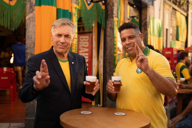 Ronaldo Fenômeno e um participante brindam com cerveja Brahma em ambiente com bandeiras, em campanha para a Copa do Mundo 2026.