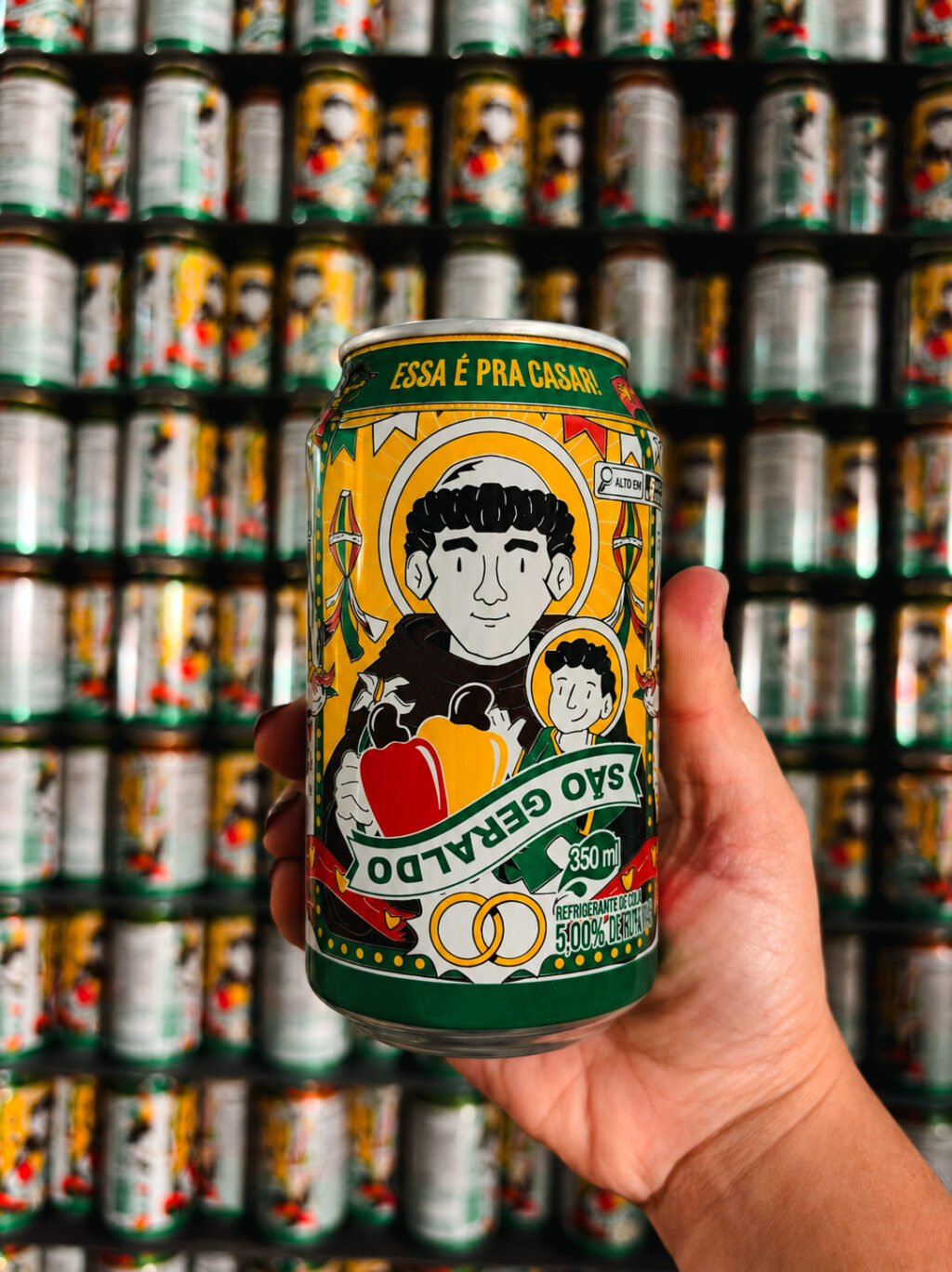 Lata de Cajuína São Geraldo com desenho junino e texto na embalagem, sendo segurada em frente a várias latas.