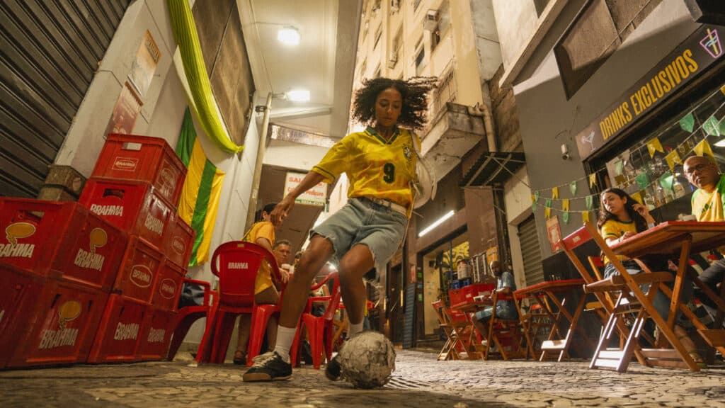 Ambiente de bar com cadeiras e mesas, caixas de cerveja Brahma e uma cena com bola ao fundo, em promoção de chopp grátis.