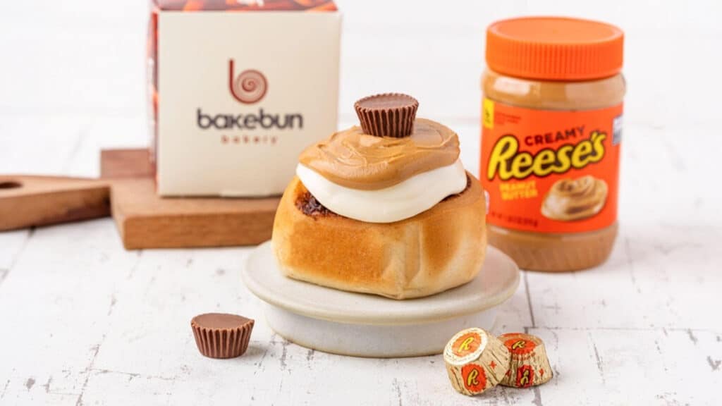 Cinnamon roll da Bakebun com cobertura e pasta de amendoim Reese’s, ao lado de potes e mini bombons Reese’s.