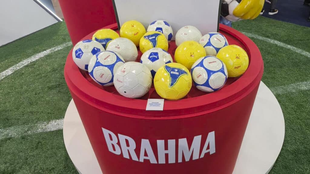 Balde vermelho com bolas colecionáveis Brahma em ambiente esportivo, com bolas brancas e amarelas