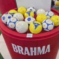 Balde vermelho com bolas colecionáveis Brahma em ambiente esportivo, com bolas brancas e amarelas
