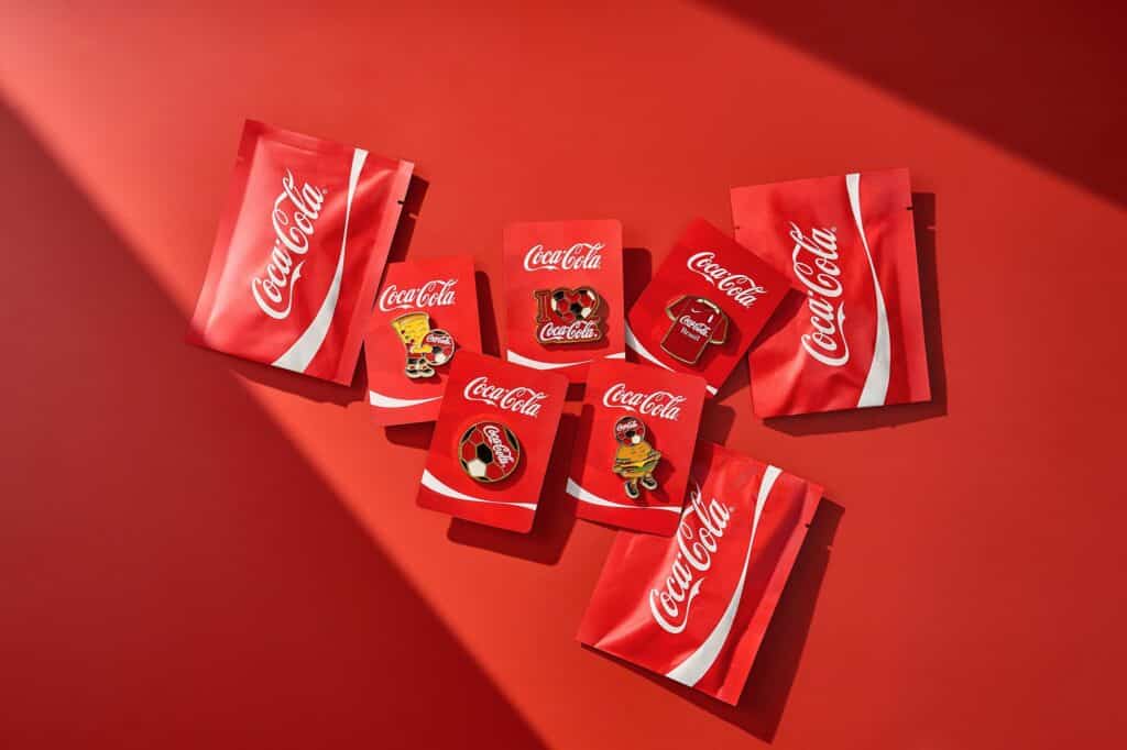 Pins colecionáveis da Coca-Cola em embalagem vermelha, com símbolos de futebol e torcida para a Copa do Mundo.