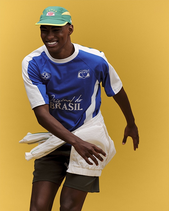 Modelo vestindo camisa azul e branca da collab Renner e Guaraná Antarctica, com boné verde, em fundo amarelo.