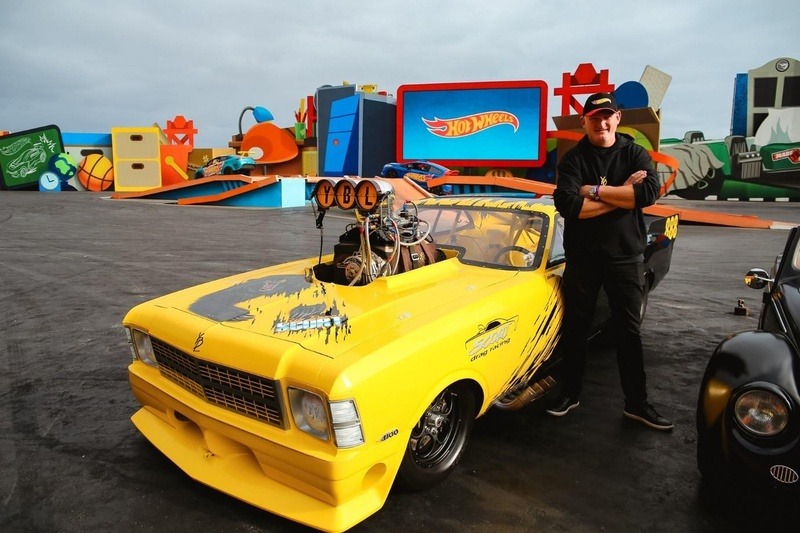 Participante posa ao lado de um carro customizado amarelo estilo Hot Wheels, em evento ao ar livre com carros e itens ao fundo.