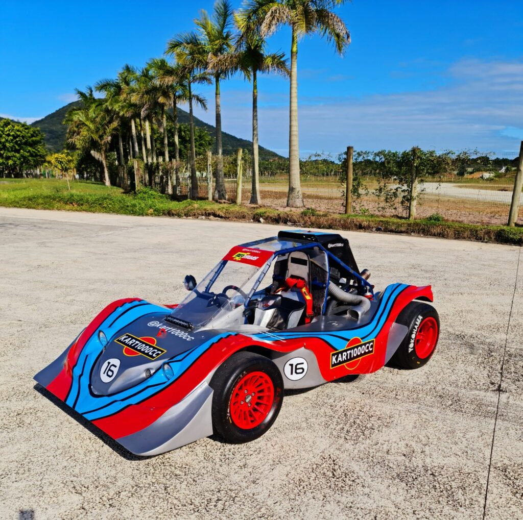 Carro customizado Hot Wheels Legends 2022, azul e vermelho, em pista de asfalto ao ar livre com palmeiras ao fundo.