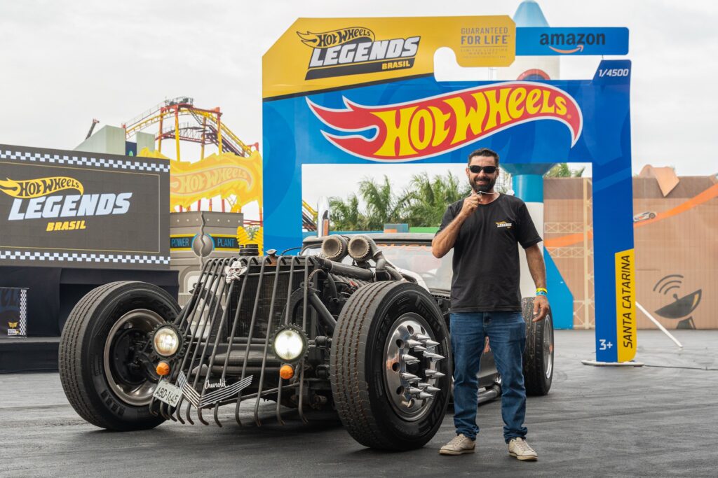 Participante ao lado de um carro customizado Hot Wheels Legends Brasil, com embalagens do concurso ao fundo