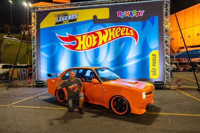 Participante posa ao lado de um carro customizado laranja no palco do Hot Wheels Legends 2024, com estande da RiHappy ao fundo.
