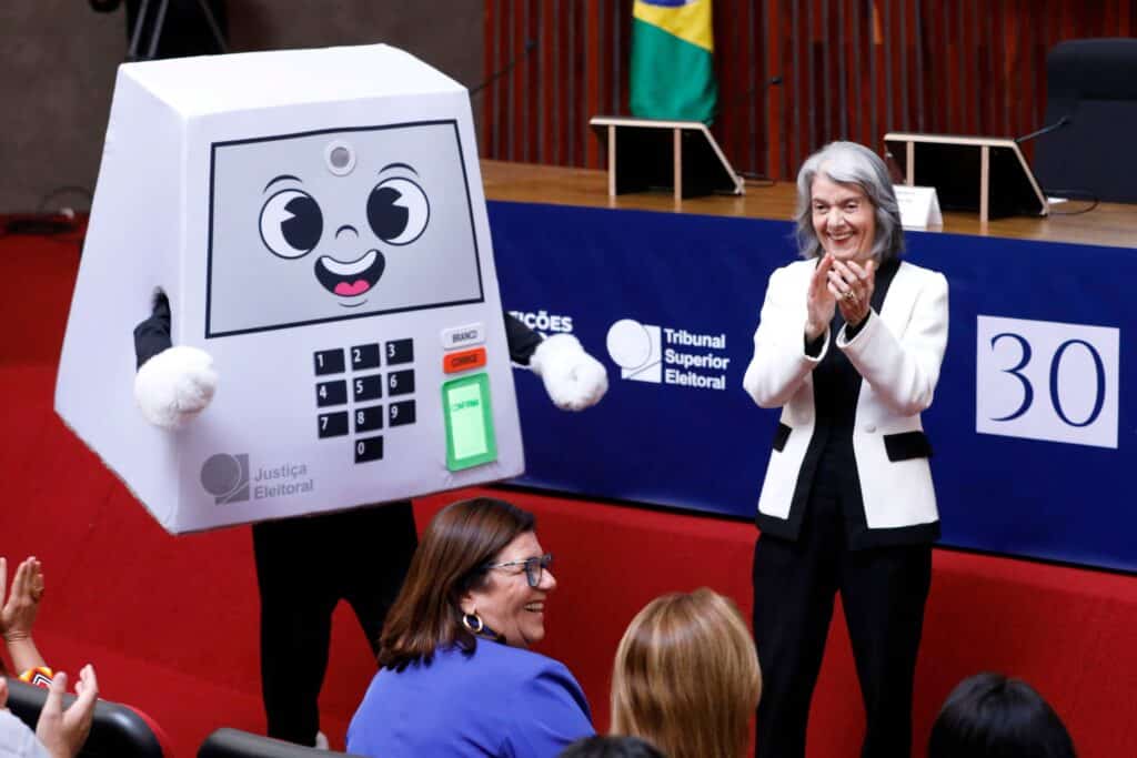 Mulher em sessão votando e apresentando urna eletrônica com rosto sorridente e telinha, ao lado de painel do tribunal eleitoral.