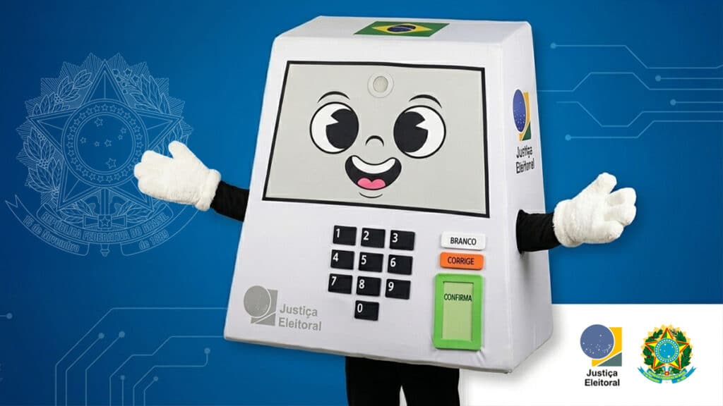 Mascote Pilili do TSE segurando uma urna eletrônica em fundo azul, com logotipos do Brasil e da Justiça Eleitoral.