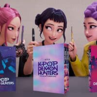 Três personagens estilo anime segurando livros de graphic novel “KPOP DEMON HUNTERS” sobre Guerreiras do K-Pop.