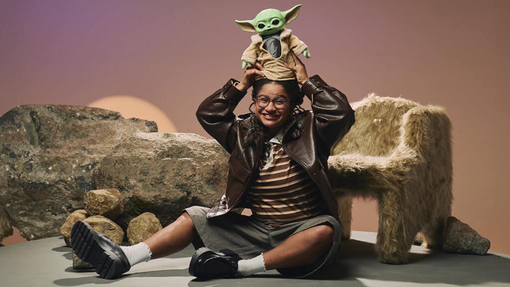 Mulher com óculos e figurino de espirito Star Wars sentada na areia, com Grogu em cima da cabeça e um felino ao lado.