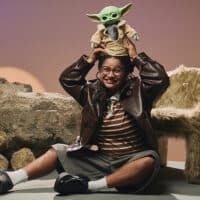 Mulher com óculos e figurino de espirito Star Wars sentada na areia, com Grogu em cima da cabeça e um felino ao lado.