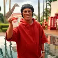 Pessoa sorridente com moletom vermelho em piscina, levantando uma lata vermelha da Itaipava em cena de campanha.