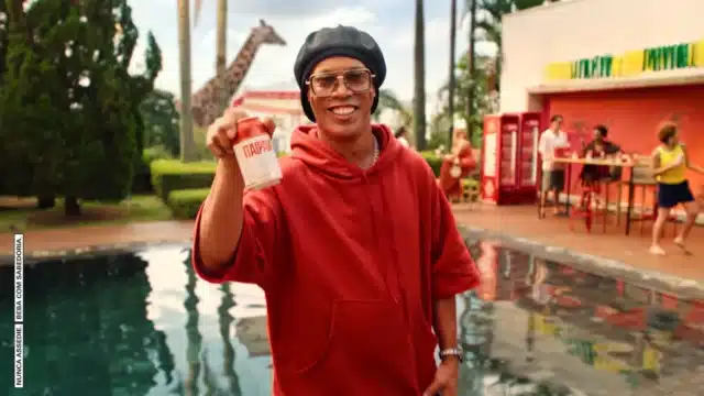 Pessoa sorridente com moletom vermelho em piscina, levantando uma lata vermelha da Itaipava em cena de campanha.