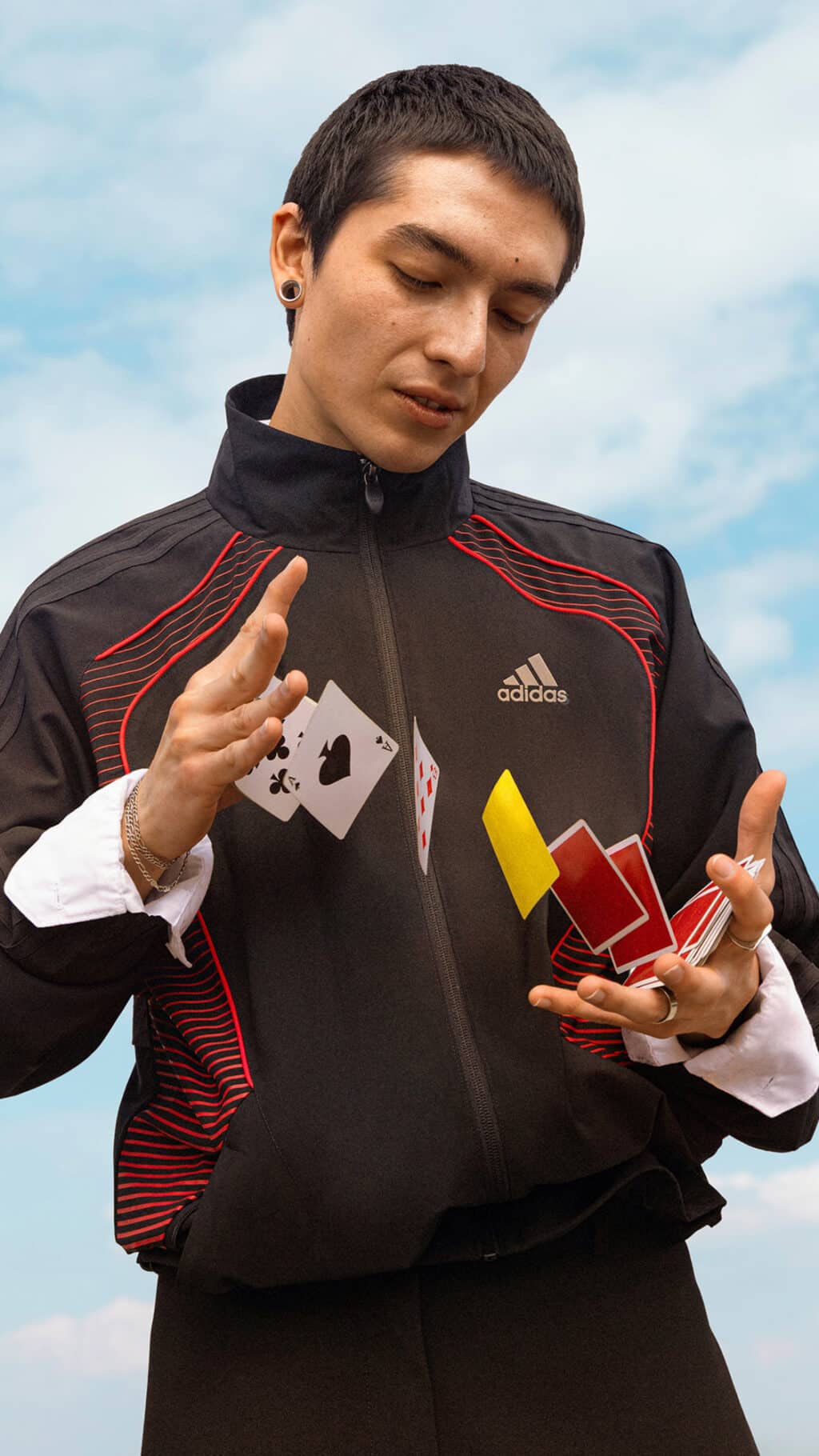 Homem segurando baralho e cartas de jogo, usando jaqueta adidas Originals esportiva, em foto ao ar livre com céu ao fundo.