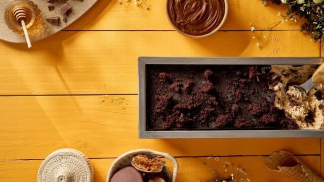 Pão de mel com cobertura e pedaços de chocolate, sobre mesa de madeira, ao lado de potes com creme de chocolate