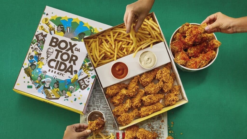 Box da Torcida KFC com frango e fritas Box da Torcida KFC com frango empanado e porção de fritas sendo compartilhado sobre fundo verde.