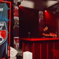 Faixa “Embaixadas Bud” em bar temático com telão ao fundo e pessoa operando equipamento em evento.