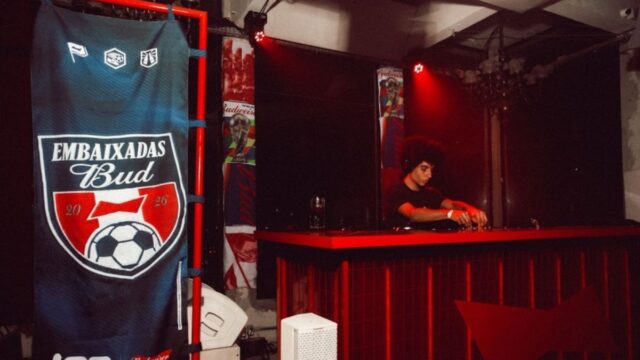 Faixa “Embaixadas Bud” em bar temático com telão ao fundo e pessoa operando equipamento em evento.