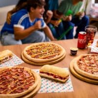 Mesa com pizzas e sanduíches da Domino’s inspirados na Copa do Mundo, com refrigerante e garrafas ao lado