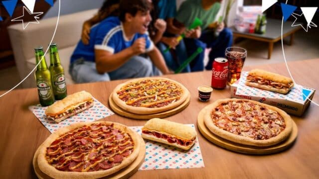 Mesa com pizzas e sanduíches da Domino’s inspirados na Copa do Mundo, com refrigerante e garrafas ao lado