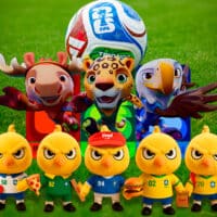 Personagens mascotes da Copa do Mundo FIFA 2026 em campo de futebol, com mascote central segurando a bola oficial