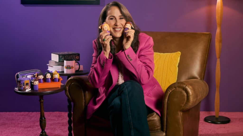 Maggie Wheeler em campanha da QDB, usando jaqueta rosa e segurando bonecas, sentada em cadeira em cenário roxo