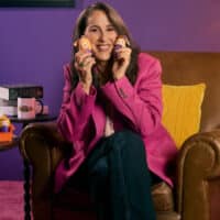Maggie Wheeler em campanha da QDB, usando jaqueta rosa e segurando bonecas, sentada em cadeira em cenário roxo