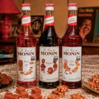 Três garrafas de xarope MONIN na mesa com doces da Festa Junina: pé de moleque, brigadeiro e doce de leite.