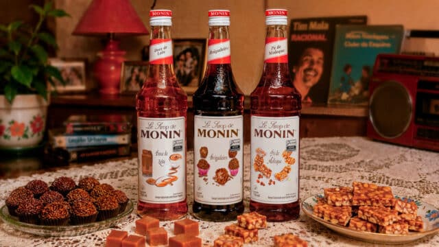 Três garrafas de xarope MONIN na mesa com doces da Festa Junina: pé de moleque, brigadeiro e doce de leite.