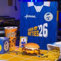 Sanduíche e batatas em embalagem da Liga Patties 2026, com caixa e copo ao lado, em cenário de Copa do Mundo.
